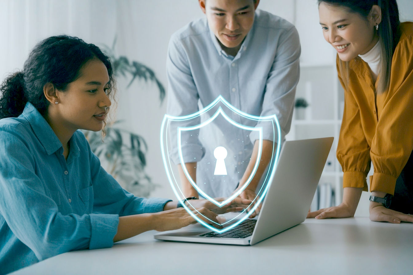 Professional, lifelike, eye-level medium shot photography of three people confidently using a sleek, modern laptop together in a bright, contemporary home office. A subtle, translucent digital shield graphic overlay hovers protectively above the laptop screen, symbolizing cybersecurity. The mood is positive, secure, and tech-savvy. Soft, natural lighting, shallow depth of field focusing on the laptop and shield. Colors are clean and modern.