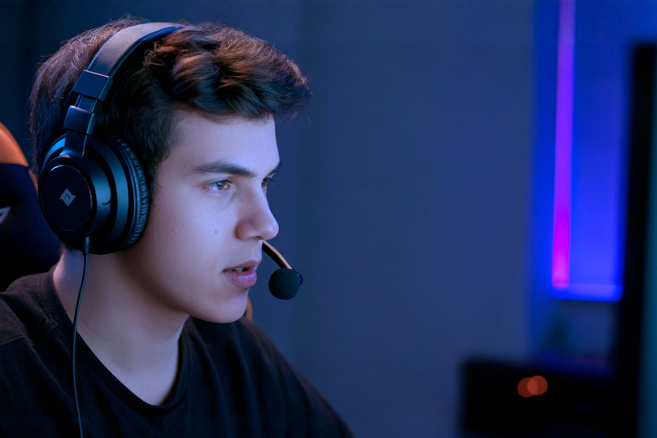 Lifelike pro photography, eye-level medium shot of a focused young adult gamer (20s) in a dimly lit but stylish gaming room with subtle RGB ambient lighting. They are wearing high-quality headphones and speaking clearly into a sleek, modern headset gaming microphone.