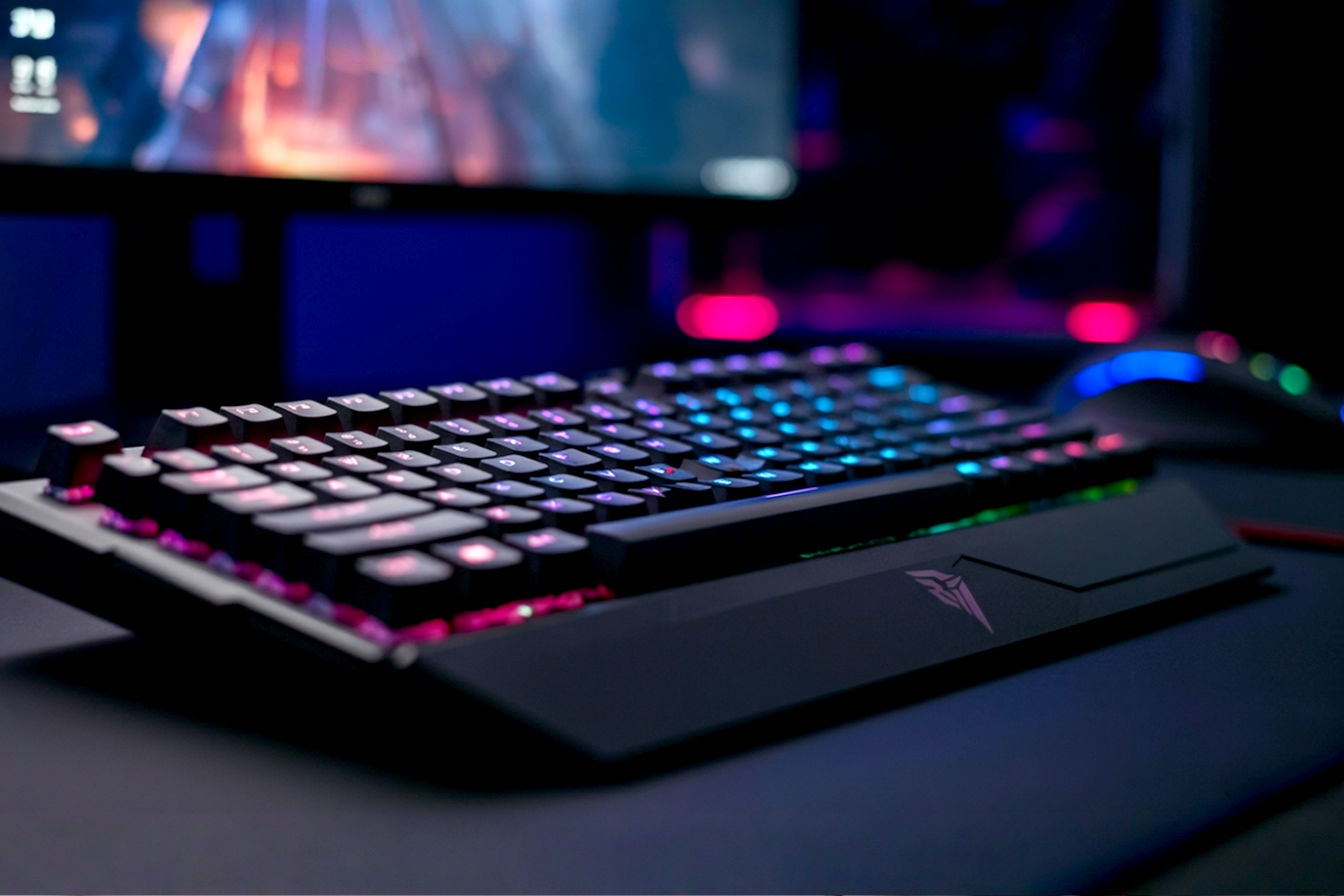 Dynamic, slightly low-angle pro photography shot of a sleek, modern mechanical gaming keyboard as the central focus. The keyboard features vibrant RGB backlighting with a subtle glow effect on the keys. One or two keycaps are mid-actuation, hinting at rapid gameplay. The background is a softly blurred gaming setup (monitor with an abstract futuristic game scene, high-end mouse). Emphasize a premium, high-performance feel.